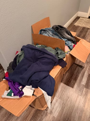 Reviewer's Premium Mix Bale with assorted clothes overflowing from an open cardboard box onto a wooden floor.