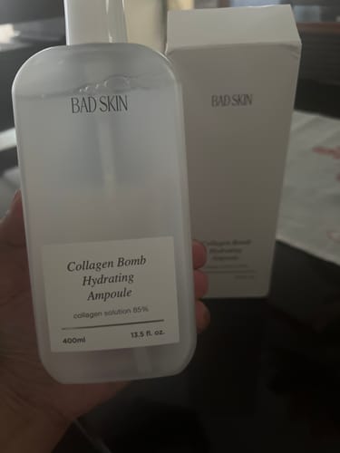 Reviewer holding the Bad Skin Collagen Bomb Hydrating Ampoule in a large, translucent bottle, with its white box in the background.