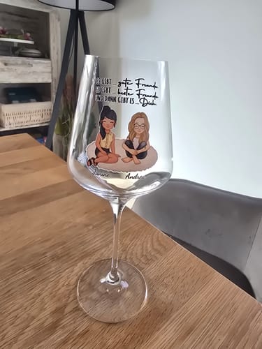 Ein Kunde präsentiert sein personalisiertes Weinglas mit einem bunten Aufdruck von zwei Freundinnen auf einem Holztisch.