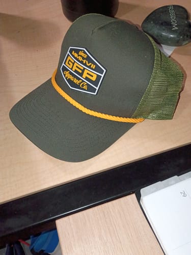 Reviewer's army green trucker hat with a yellow rope and a GFP Apparel Co. patch on a desk.