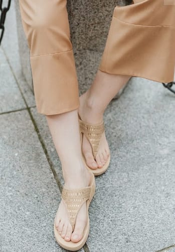 A customer's feet wearing the beige Drift sandals with studded T-straps while sitting on a stone surface.