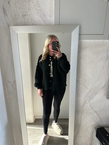 Kundin trägt den schwarzen Oversized-Cardigan "CozyGlam" in einer Ganzkörperansicht im Spiegel.
