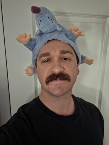 Reviewer taking a selfie while wearing the light blue plush 'Lester's Possum Pal' hat.