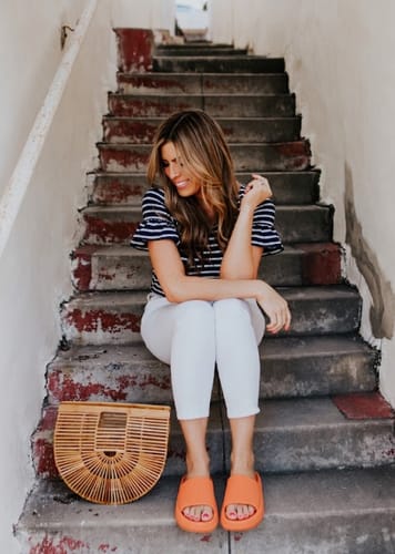 Customer sitting on stairs wearing bright orange Cloud Feet slides with white pants.
