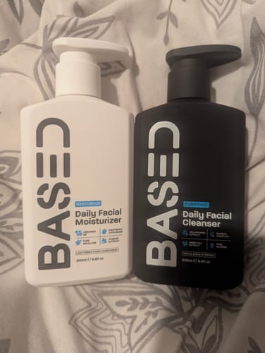 Customer's Daily Skincare Duo, with the white moisturizer bottle and black cleanser bottle sitting on a bed.