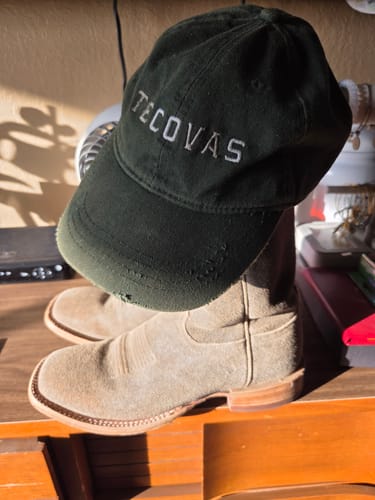 Reviewer's suede The Doc Western Boots on a wooden surface with a Tecovas cap resting on the boot shaft.