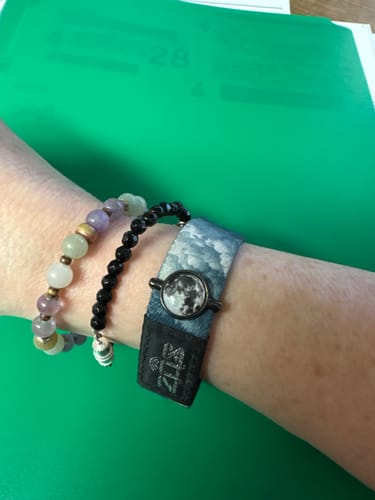 Reviewer's wrist showing the round Moon Charm attached to a fabric bracelet with a cloud design, next to other beaded bracelets.