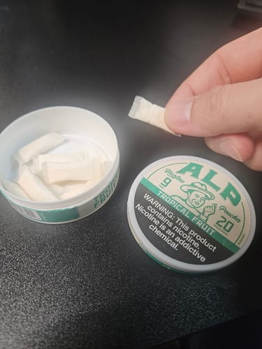 Customer holds a soft ALP nicotine pouch. The open tin of 'Tropical Fruit' flavor pouches is on the desk.