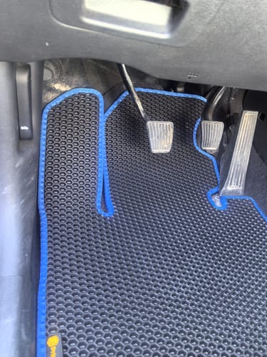 Recenzent pokazuje czarny dywanik samochodowy o wzorze plastra miodu z niebieskim obszyciem, dopasowany do podłogi w aucie.