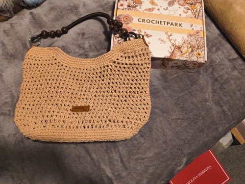 Reviewer's completed beige crocheted bag, featuring a custom leather label on the front, sits next to its box.