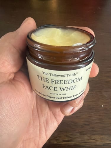 A customer holds an open amber jar of Freedom Face Whip, showing the pale yellow cream inside.