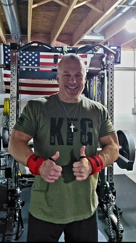 Reviewer smiling in a home gym while wearing the green KFG t-shirt and giving two thumbs-up.