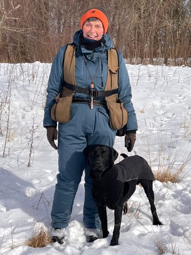 Anmelder i en blå heldragt, der står med sin hund i et snedækket landskab.