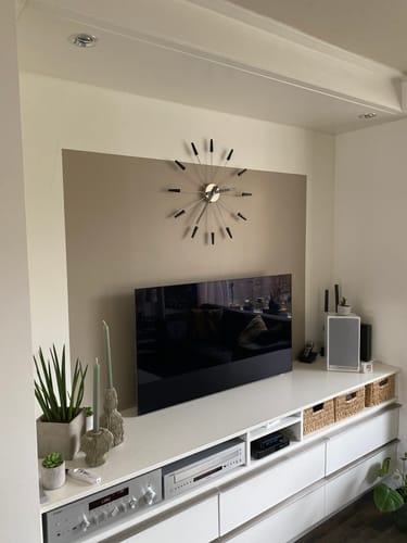 Klant toont de NeXtime Plug Inn wandklok aan de muur boven een tv in een moderne woonkamer.