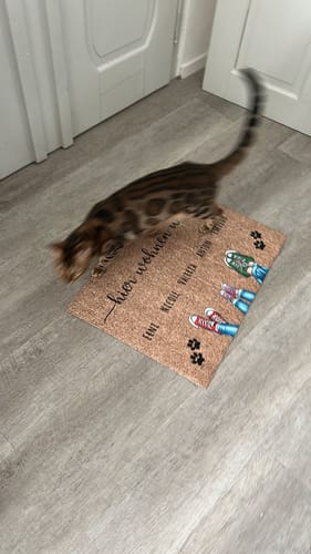 Kundenbewertung: Eine gefleckte Katze läuft über die personalisierte 'Schuhe und Pfoten'-Fußmatte, die auf einem Holzboden liegt.
