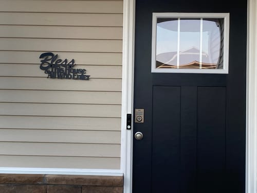 Customer's black metal Bless This House Decor mounted on the siding of a house next to the front door.