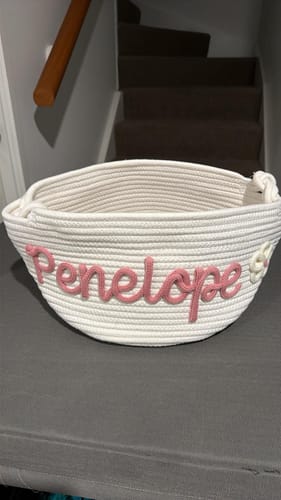 Customer's personalized white knitted basket with the name 'Penelope' in pink yarn and a flower icon.