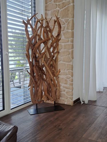 Ein Kunde zeigt den Teak Holz Raumteiler RANTING, der in einem Wohnzimmer vor einer Steinwand aufgestellt ist.