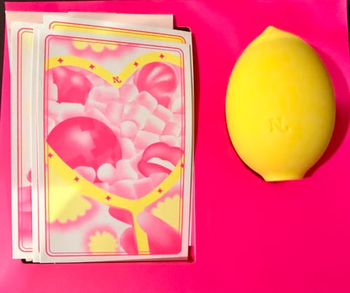 Reviewer's yellow Lem Clitoral Massager on a bright pink surface, shown with its illustrated packaging cards.