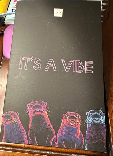 Customer's March 2026 Booklet featuring a black cover with 'IT'S A VIBE' in pink text and four colorful otters at the bottom.