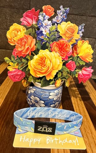 Customer's Happy Birthday Bracelet on its display card, set in front of a colorful pop-up paper flower bouquet.