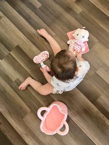 Overhead view of a baby on a wood floor playing with a Personalized Pudding Baby Girl doll and its pink accessories.