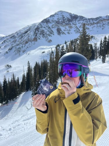 Customer in ski gear eating a Cael's Cookie on a snowy mountain slope.