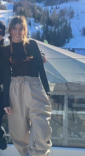 Reviewer smiling while wearing baggy beige snow pants in a snowy mountain setting.