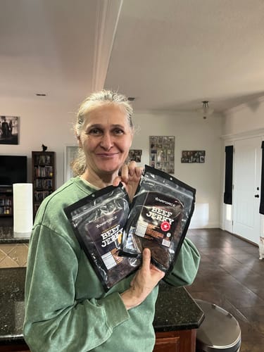 Reviewer smiling while holding two bags of Gourmet Premium Beef Jerky in a kitchen.