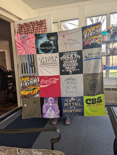 Customer holding up their large, colorful Premium T Shirt Quilt made from various t-shirts in a sunroom.