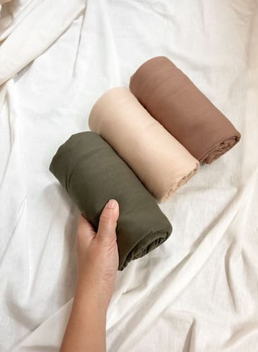 Customer's hand holding a rolled Lovemère Buttery Nursing Cover in Matcha, with the Latte and Mocha colors in the background.