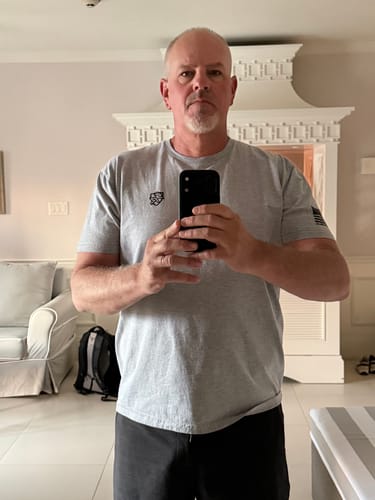 Customer shows the fit of their grey Shield Branded Tee, with snug arms, a looser stomach, and a flag patch on the sleeve.