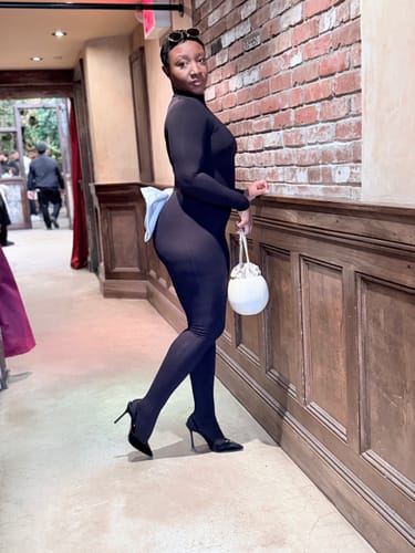 Customer poses in the black, form-fitting Naked Closed Toe Bow Jumpsuit with a light blue back bow.