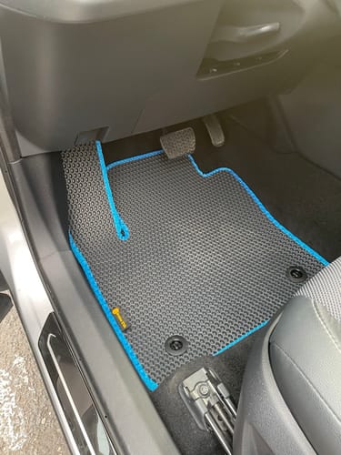Recenzent pokazuje czarny dywanik samochodowy z niebieską lamówką, dopasowany do podłogi po stronie kierowcy.