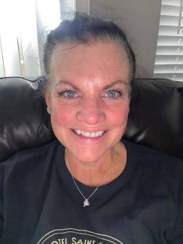 A smiling reviewer wears their silver Redeemed Necklace with a black shirt while sitting on a couch.