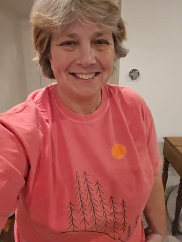 A smiling reviewer wearing the coral Sunrise Pines T-shirt, showing the fit and graphic detail.