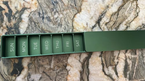 Customer's green Weekly Case open on a counter, showing the seven daily compartments labeled SUN through SAT.