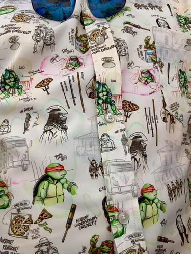Close-up of the reviewer's 'Heroes in a Half Sketch' shirt, showing the detailed pattern of TMNT character sketches and icons.