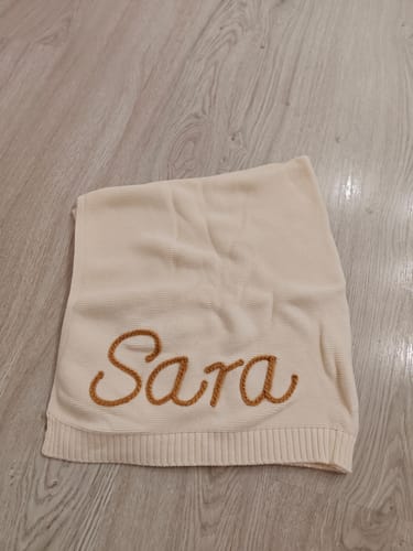 Customer's personalized hand-knitted blanket in cream, with the name 'Sara' embroidered in brown yarn.