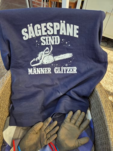 Rezensent zeigt das dunkelblaue MÄNNERGLITZER KETTENSÄGE Sweatshirt, das auf einem Stuhl mit Arbeitshandschuhen liegt.