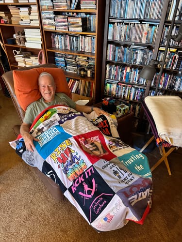Customer smiling in a recliner while covered with their colorful Premium T Shirt Quilt.