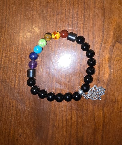 A customer's crystal bracelet with colorful chakra beads, black beads, and a silver Hamsa charm on a wooden surface.