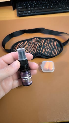 Reviewer holding a bottle of Melatonin Sleep Spray, with a sleep mask and earplugs on a desk in the background.