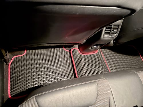 Recenzent pokazuje dopasowane dywaniki samochodowe z czerwonymi obszyciami, zainstalowane na tylnej podłodze samochodu.