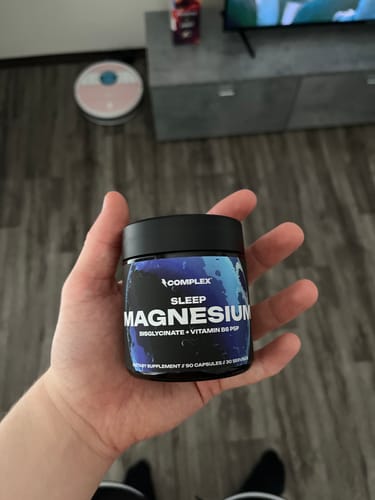 A customer's hand holding a black jar of Sleep Magnesium supplement in a living room setting.