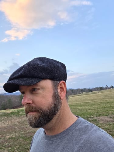 Reviewer outdoors wearing the dark tweed JP Cap, shown in profile against a blue sky.