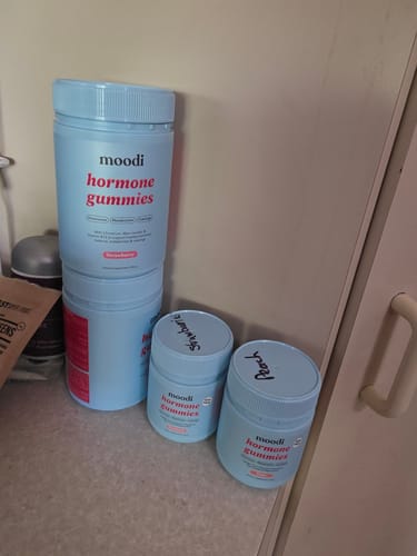 Customer's four light blue bottles of Moodi Hormone Gummies on a counter, with 'Strawberry' and 'Peach' handwritten on two of the lids.