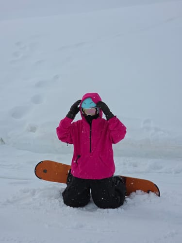 Reviewer kneeling in the snow, wearing a bright pink Half-Zip Snow Anorak with a snowboard nearby.