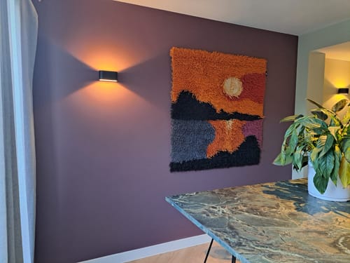 Klant toont het kleurrijke HKliving Wandkleed in de avond aan een paarse muur, naast een wandlamp.