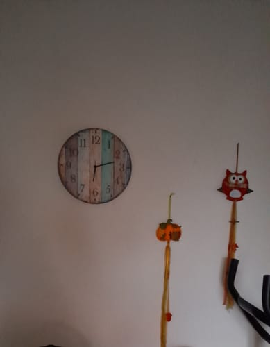 Kunde zeigt die Wanduhr aus Holz Vintage Summer, die an einer schlichten Wand hängt.